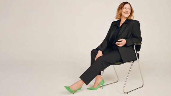 A woman in a black suit with green heels sitting in a chair against a light background. She is looking over her shoulder and smiling.