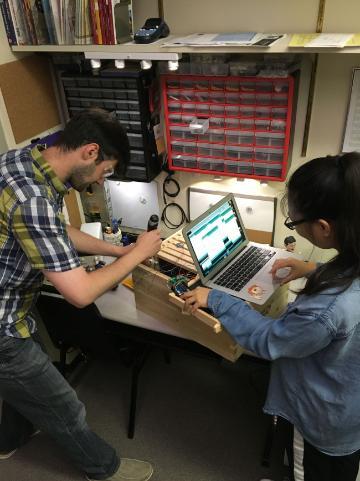 Two people assembling hardware on a desk, surrounded by tools and electronic equipment.