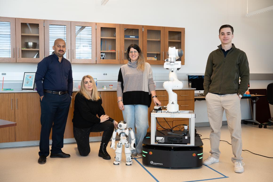 Photo of 3 PHD students and faculty Maria Kyrarini posing with a small knew-height robot and a 5-foot tall robotic arm.