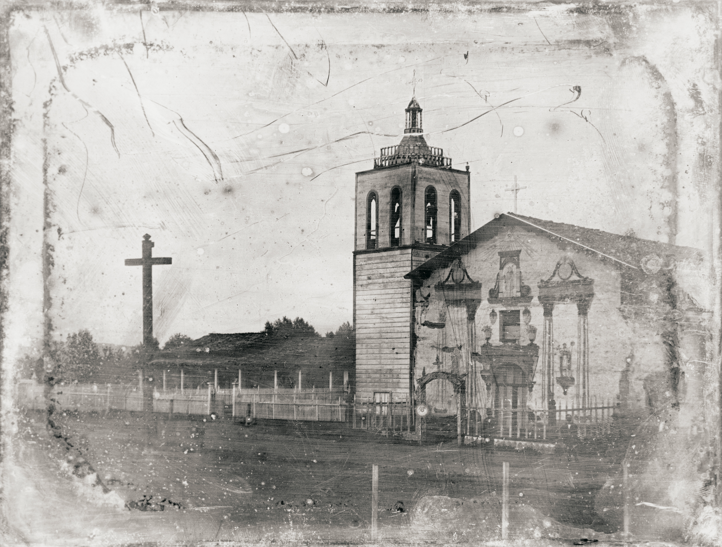 Black and white photograph of an old mission church with a cross nearby.
