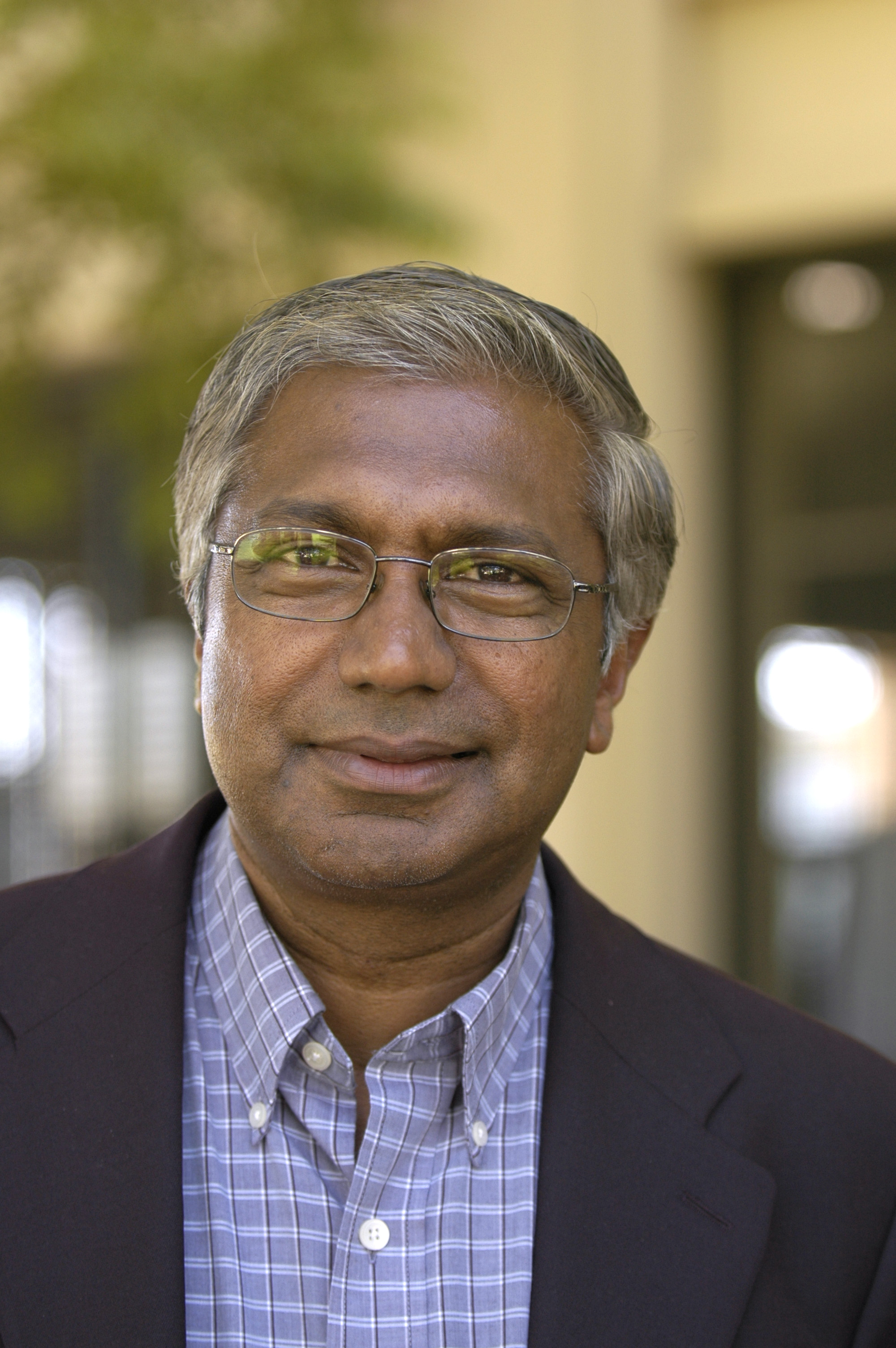 A man wearing glasses, a suit jacket, and a collared shirt, standing outdoors.