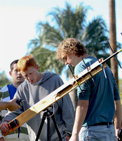People examining a telescope outdoors with palm trees in the background.