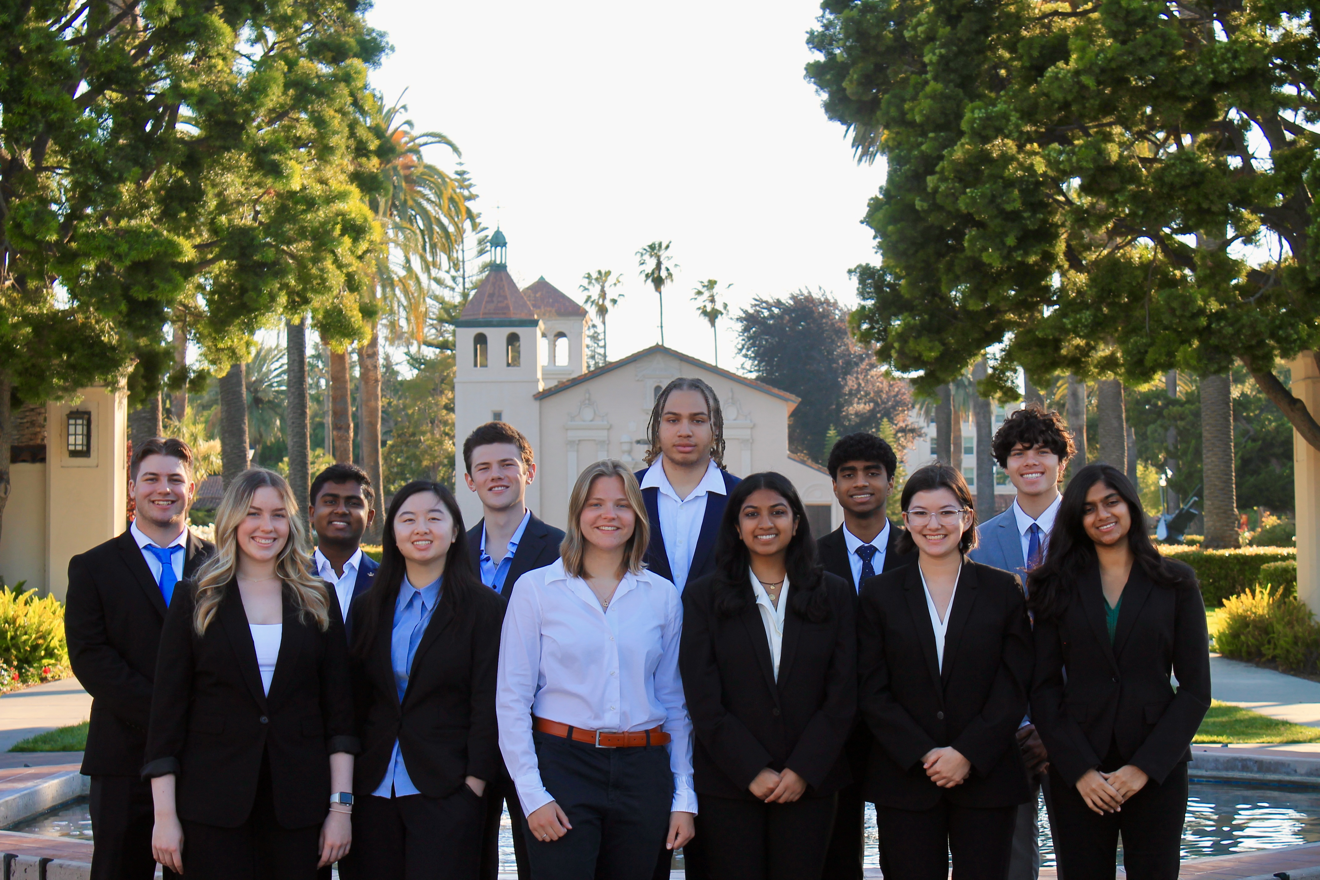 Group photo of 12 students in professional attire in front of the Santa Clara Mission.