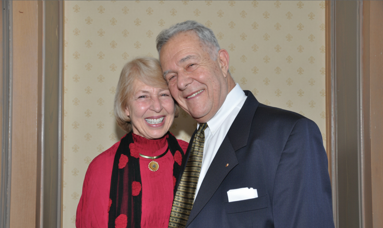 A happy elderly couple, dressed in formal attire, smiles warmly at the camera, with the woman in red and the man in a dark suit.