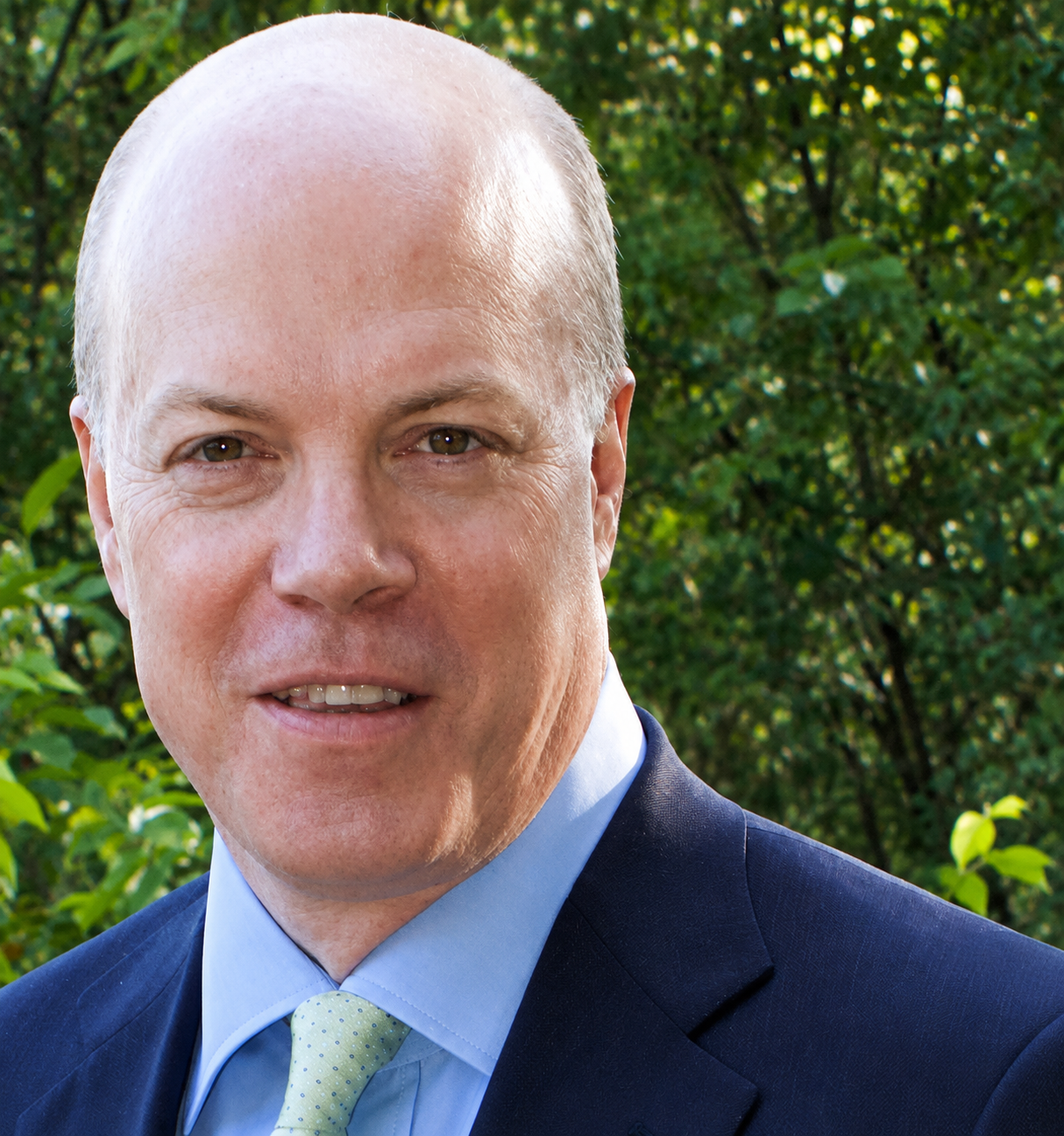 A headshot of Prof. Stephen Diamond a white man with a shaved head, wearing a light blue shirt, lime green tie and navy suit jacket the background is greenery