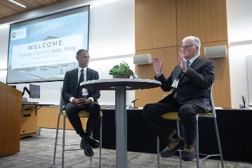 Two guys at a round table with a mic in one of guys hand speaking to a crowd