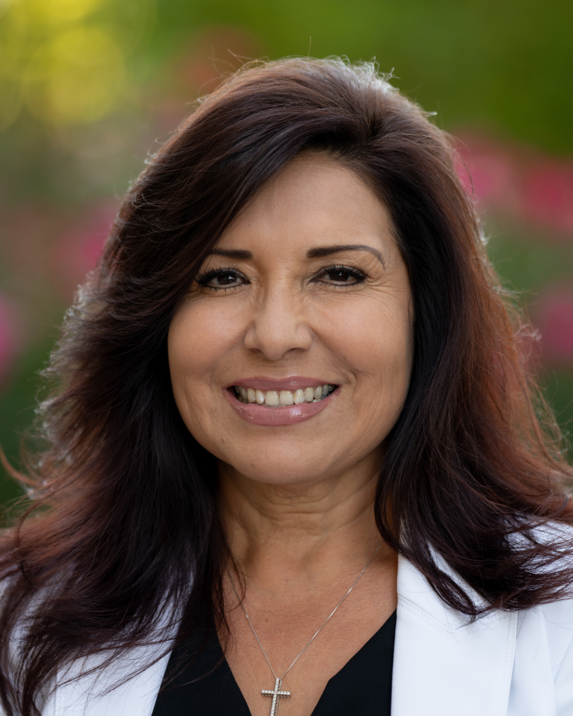 Headshot of a smiling woman with long dark hair wearing a white blazer and cross necklace against a blurred outdoor background.