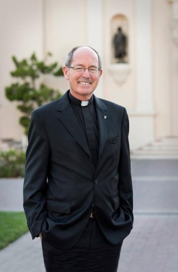 A man in a black clergy attire standing outdoors, smiling.